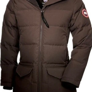 Brun dunjacka från Canada Goose - Säljer en brun dunjacka från Canada Goose med dragkedja och lång ärm. Jackan har en klassisk design med två stora fickor framtill och en logotyp på ärmen. Perfekt för kalla dagar.