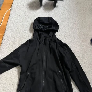 Svart Cp company hoodie - Säljer en svart jacka från C.P. Company med unik design. Jackan har en huva med inbyggda glasögon och dragkedja framtill. Perfekt för en cool och modern stil.