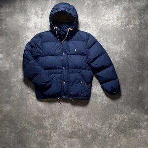 Ralph Lauren - Säljer en snygg mörkblå dunjacka från Ralph Lauren med huva och dragkedja. Jackan har knappar och en broderad logga på bröstet. Perfekt för kyliga dagar! Säljer då den är lite för kort. Storlek L men passar även M