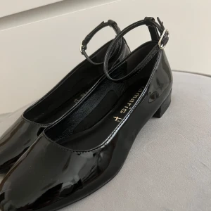 Svarta lackade ballerinaskor från Tamaris - Säljer ett par snygga svarta ballerinaskor från Tamaris i storlek 35 som är endast använd en gång. De är i lackat material med en elegant rem runt ankeln som har ett spänne i silver. Perfekta för både vardag och fest!