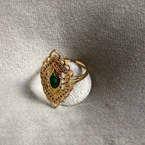 Guldfärgad ring med grön sten - Säljer en superfin guldfärgad ring med en vacker grön sten i mitten. Ringen har ett intrikat mönster som ger den en elegant och unik look. Perfekt för att piffa upp vilken outfit som helst! Sjukt bra kvalitet. Har inte tappat färgen så länge jag har haft den, inga tecken på användning!