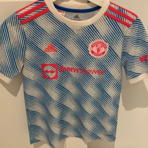 Manchester United fotbollströja från Adidas - Säljer en cool Manchester United fotbollströja från Adidas. Tröjan är vit och blå med ett randigt mönster och har korta ärmar. Den har röda detaljer och klubbens emblem på bröstet. Perfekt för unga fans som vill visa sitt stöd!