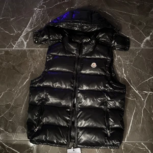 Svart dunväst från Moncler - Snygg svart dunväst från Moncler med dragkedja och huva. Västen har en glansig finish och två praktiska fickor med dragkedjor. Perfekt för kyligare dagar när du vill hålla stilen👌🏻priset ej hugget i sten. Var inte rädd för att höra av er😉storlek M men passar L