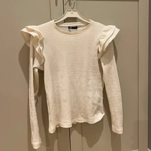 Vit ribbad tröja från Zara - Säljer en stilren vit ribbad tröja från Zara med volangdetaljer på axlarna. Tröjan har långa ärmar och en rund halsringning. Är storlek S och säljs inte längre💌