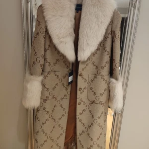 Beige kappa med pälsdetaljer från Gant - Elegant beige kappa från Gant med lyxiga pälsdetaljer på krage och ärmslut. Kappan har ett diskret mönster med Gant-logotypen och knappar framtill. Perfekt för att hålla stilen under kyliga dagar.