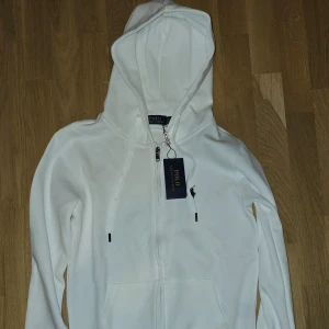 Vit hoodie från Ralph Lauren - Säljer en helt ny stilren vit hoodie från Ralph Lauren med dragkedja och klassisk logga på bröstet. Perfekt för en avslappnad look. Huvan har dragsko för justerbar passform. Stl M