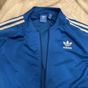Blå träningsjacka från Adidas - Säljer en klassisk blå träningsjacka från Adidas med vita ränder längs ärmarna och det ikoniska trefoil-logotypen på bröstet. Jackan har dragkedja framtill och en sportig look.
