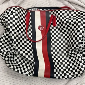 Rutig shoppingväska från Tommy Hilfiger - Snygg shoppingväska från Tommy Hilfiger med ett svartvitt rutigt mönster och röda detaljer. Väskan har en dragkedja och ett stilrent randigt band i rött, vitt och svart på framsidan. Perfekt för en trendig look!