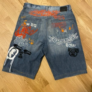 Blå jeansshorts med graffiti-mönster - Coola blå jeansshorts från Ecko Unltd med unikt graffiti-mönster i orange och vitt. Shorts med baggy fit och flera detaljer som broderier och tryck. Perfekta för en avslappnad stil.