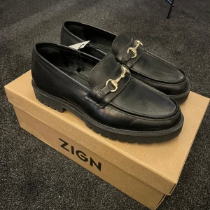 Svarta loafers från Zign - Snygga svarta loafers från Zign med gulddetaljer på ovansidan. Säljer dessa helt nya skor frän Zign! Storlek 38 men passar även 39. Inga defekter! Box tillkommer.