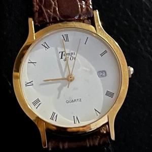 Temps d'Or herrklocka med brun läderrem - Elegant herrklocka från Temps d'Or med en klassisk vit urtavla och romerska siffror. Klockan har en gyllene boett och en datumvisare. Den bruna remmen är gjord av äkta läder med krokodilpräglat mönster.