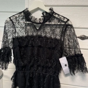 !RESERVERAD! Svart spetsblus från bubbleroom  - Elegant svart spetsblus från bubbleroom med hög hals och transparenta detaljer. Blusen har trekvartsärmar och en vacker volangdesign. Perfekt för en stilfull kväll. (OBS knappen på baksidan har gått sönder men ingenting man ser när på)