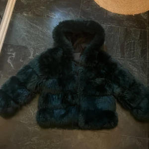 Mörkgrön pälsjacka - Säljer en lyxig mörkgrön pälsjacka med fluffig textur. Jackan har en mysig huva och långa ärmar, perfekt för kyliga dagar. Den är både stilren och trendig, vilket gör den till ett måste i garderoben. Den går att knäppa!