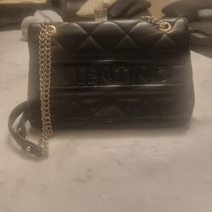 Svart quiltad axelväska från Valentino - Snygg svart axelväska från Valentino med quiltad design och kedjerem. Väskan har en elegant look med märkets logga på framsidan. Perfekt för att ge en lyxig touch till din outfit.