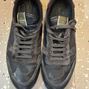 Valentino Garavani sneakers i camouflage - Snygga Valentino Garavani sneakers med ett coolt camouflage-mönster. Skorna har snörning och är i en mörk färgskala med inslag av grått och svart. Perfekta för en stilren och modern look. Äkta Valentino Rock Runners, begagnad. Priset kan diskuteras, Storlek 44 men passar även 45