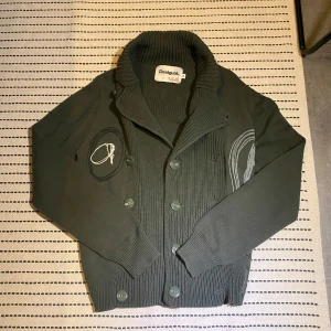 Grön kofta från Desigual - Snygg grå kofta från Desigual med unika broderade detaljer och knappar framtill. Den har en ribbad krage och muddar, vilket ger en stilren look. Perfekt för kyligare dagar. Kan skicka bild med plagget på. (Pris kan diskuteras)