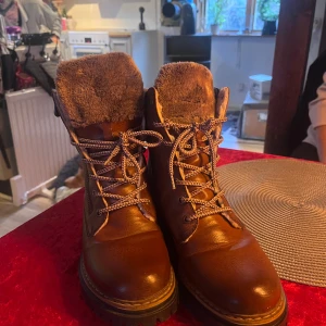 Bruna skinnboots med foder. Nypris 950kr - Säljer ett par bruna skinnboots från Rieker med snörning och dragkedja. De har ett varmt foder och en robust sula, perfekt för kyligare väder.