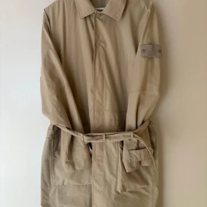 Beige trenchcoat med bälte - Snygg beige trenchcoat med klassisk design och bälte i midjan. Jackan har långa ärmar och knappar framtill. Perfekt för en stilren look.