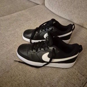 Svarta Nike sneakers med vit sula - Snygga svarta Nike sneakers med vit sula och vit swoosh-logga. Skorna har snörning och en klassisk design som passar perfekt till vardags. De är tillverkade i syntetmaterial och har en bekväm passform.