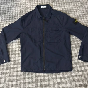 Mörkblå jacka från Stone Island - Snygg mörkblå jacka från Stone Island med dragkedja och två bröstfickor. Jackan har långa ärmar och en klassisk krage. Perfekt för en stilren look.