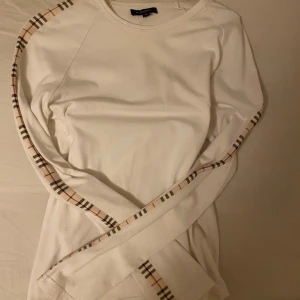 Vit långärmad tröja från Burberry - Säljer en stilren vit långärmad tröja från Burberry med klassiska rutiga detaljer längs ärmarna. Tröjan är i storlek M och har en mjuk och bekväm passform.