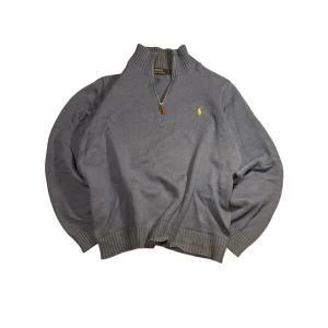 Grå tröja med dragkedja från Polo Ralph Lauren - Snygg grå tröja från Polo Ralph Lauren med halv dragkedja och ribbad krage. Den har en gul broderad logotyp på bröstet och ribbade muddar vid ärmslut och nederkant. Perfekt för en stilren look.