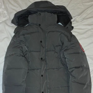 Mörkgrå dunjacka från Canada Goose Stl. M - Säljer en helt ny mörkgrå dunjacka från Canada Goose med avtagbar pälskant på huvan. Jackan har en dragkedja och knappar framtill samt en broderad logotyp på ärmen. Perfekt för kalla vinterdagar.