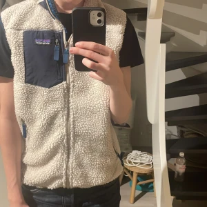 Patagonia väst - Säljer en beige fleeceväst från Patagonia med marinblå detaljer. Västen har en dragkedja framtill och en praktisk bröstficka med dragkedja. Perfekt för lager-på-lager under kyliga dagar. Storlek L men passar M