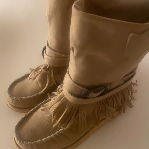 Beige mocka boots med fransar - stl 36 - Snygga beige boots i mocka med fransdetaljer runt skaftet. De har en unik design med dekorativa sömmar och en bred rem över vristen. Perfekta för en bohemisk stil.