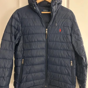 Blå pufferjacka från Polo Ralph Lauren - Säljer en snygg blå pufferjacka från Polo Ralph Lauren med dragkedja och huva. Jackan har långa ärmar och två praktiska fickor med dragkedja.