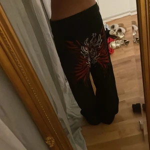 Svarta byxor med röd och vit broderad design från Shein - Snygga svarta byxor från Shein med en cool röd och vit broderad design på framsidan. Byxorna har en hög midja och är tillverkade i 100% bomull. Perfekta för att ge din outfit en unik touch.