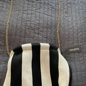 Randig axelväska med kedja från Bobobo - Snygg axelväska från Bobobo med svart och vit randigt mönster. Väskan har en elegant guldfärgad kedja som axelrem och är perfekt för att ge en stilren touch till din outfit.