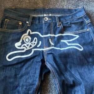 BBC Ice Cream Jeans - Storlek ca W29/L30. Coola jeans från Ice Cream med ett unikt tryck av en tecknad figur på framsidan och dödskallemotiv på bakfickorna. Mörkblå denim med kontrastsömmar och knappstängning. Perfekta för dig som vill sticka ut! Pris kan diskuteras! 