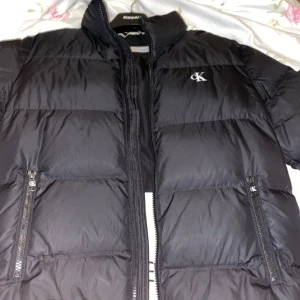 Svart pufferjacka från Calvin Klein - Snygg svart pufferjacka från Calvin Klein med dragkedja och logotyp på bröstet och ärmen. Storlek XL men jag har M/L och den sitter perfekt på mig. Jackan har praktiska fickor med dragkedja. Bara höra av sig vid frågor och funderingar🫡🥶