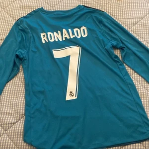 Blå Real Madrid fotbollströja med Ronaldo 7 - Snygg långärmad Real Madrid tröja i blått med Ronaldo 7 på ryggen. Tröjan har Adidas-logga och Fly Emirates som sponsortryck på framsidan. Perfekt för fotbollsfans!