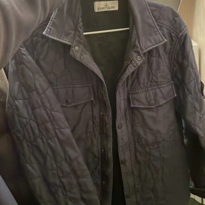 Svart quiltad jacka från Stone Island - Säljer en blå quiltad jacka från Stone Island med klassisk krage och knappar framtill. Jackan har två bröstfickor med lock och den ikoniska logotypen på ärmen. Perfekt för en stilren look.