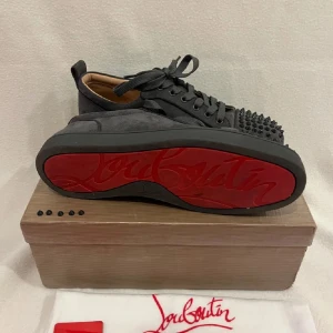 Grå sneakers med nitar från Louboutin - Snygga grå sneakers från Louboutin med nitar på tån och snörning. Skorna har en ikonisk röd sula och är tillverkade i mocka. Perfekta för att ge din outfit en edgy touch.