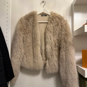 Beige pälsjacka från Gina - Säljer en fluffig och trendig beige pälsjacka från Gina. Jackan har en öppen design utan knappar eller dragkedja och är perfekt för att hålla sig varm och stilren under kyliga dagar. Använd 1 gång