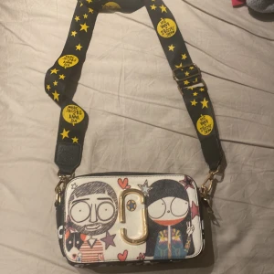 Färgglad axelväska från Marc Jacobs - Säljer en limeted edition axelväska från Marc Jacobs. Väskan har en justerbar axelrem med gula stjärnor och text. Den har en lila kant och dragkedja i guld. Perfekt för att ge din outfit en rolig touch! Fick den av min moster och hon sa att den va riktig men jag har inge bevis skriv vid mer frågor