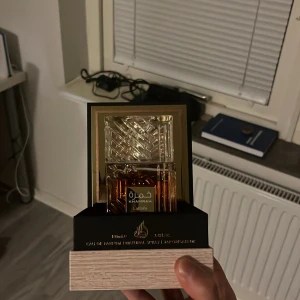 Khamrah Eau de Parfum - Elegant parfym från Lattafa, Khamrah, med en lyxig flaska i guld och brunt glas. Flaskan har ett intrikat mönster på korken och en stilren design. Perfekt för den som söker en sofistikerad doftupplevelse.
