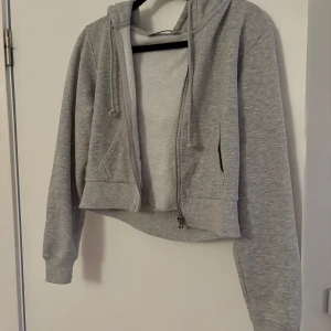 Grå croppad hoodie från Bershka - Säljer en grå croppad hoodie från Bershka med dragkedja och huva. Perfekt för en avslappnad stil. Den har långa ärmar och ribbade muddar.