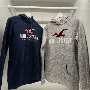 Hoodies från Hollister - Två hoodies från Hollister i olika färger. Den ena är marinblå med rosa och vit logga, och den andra är gråmelerad med röd och vit logga. Båda har huva och känguruficka, perfekt för en avslappnad stil. 100 kr styck. 
