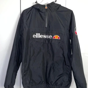 Svart vindjacka från Ellesse - Snygg svart vindjacka från Ellesse med dragkedja och stort logotryck på bröstet. Jackan har en huva och långa ärmar, perfekt för blåsiga dagar. Säljer den för att jag inte använder . Jackan är använd ett antal gånger men i mkt bra skick. Storlek är xs men passar också S