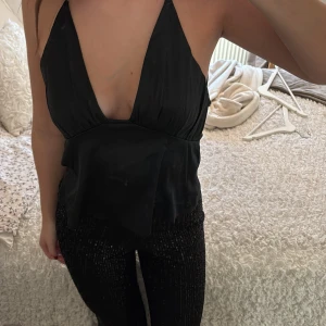 Svart halterneck linne - Snyggt svart halterneck linne med djup urringning och smala axelband som korsas i ryggen. Perfekt för en kväll ute eller en festlig tillställning.