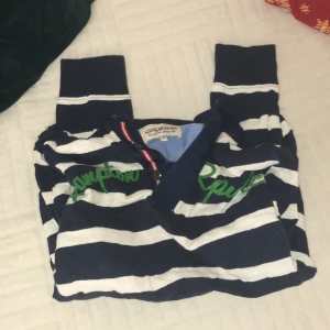 Randig tröja från Hampton Republic - Säljer en snygg randig tröja från Hampton Republic i blått och vitt. Tröjan har gröna detaljer och en krage med knappar . Perfekt för en avslappnad stil.