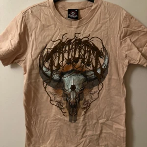 Beige t-shirt från Tribal - Säljer en beige t-shirt från Tribal Gear med ett stort tryck av en dödskalle och horn på framsidan. T-shirten är tillverkad i 100% bomull och har en normal passform med korta ärmar. Perfekt för en avslappnad stil.