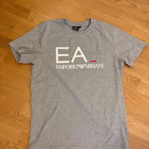  Emporio Armani T-shirt  - Snygg grå t-shirt från Emporio Armani med tryck av logotypen 'EA' och 'Emporio Armani' i vitt och rött på framsidan. Perfekt för en stilren och avslappnad look.