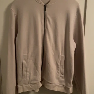 Beige tröja från Bläck - Snygg beige tröja från Bläck med dragkedja framtill. Tröjan har en stilren design med långa ärmar och ribbade muddar vid ärmslut och krage. Perfekt för en avslappnad look.