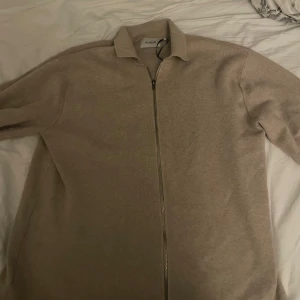 Beige tröja med dragkedja  - Säljer en stilren beige tröja från SNOOT med dragkedja framtill. Perfekt för en avslappnad look. Tröjan har långa ärmar och en klassisk krage. Passar bra till både jeans och chinos.