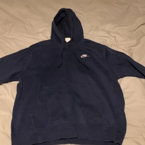 Mörkblå hoodie från Nike - Säljer en mörkblå hoodie från Nike med klassisk logga på bröstet. Tröjan har en stor ficka framtill och justerbar huva med snören. Perfekt för en avslappnad stil.